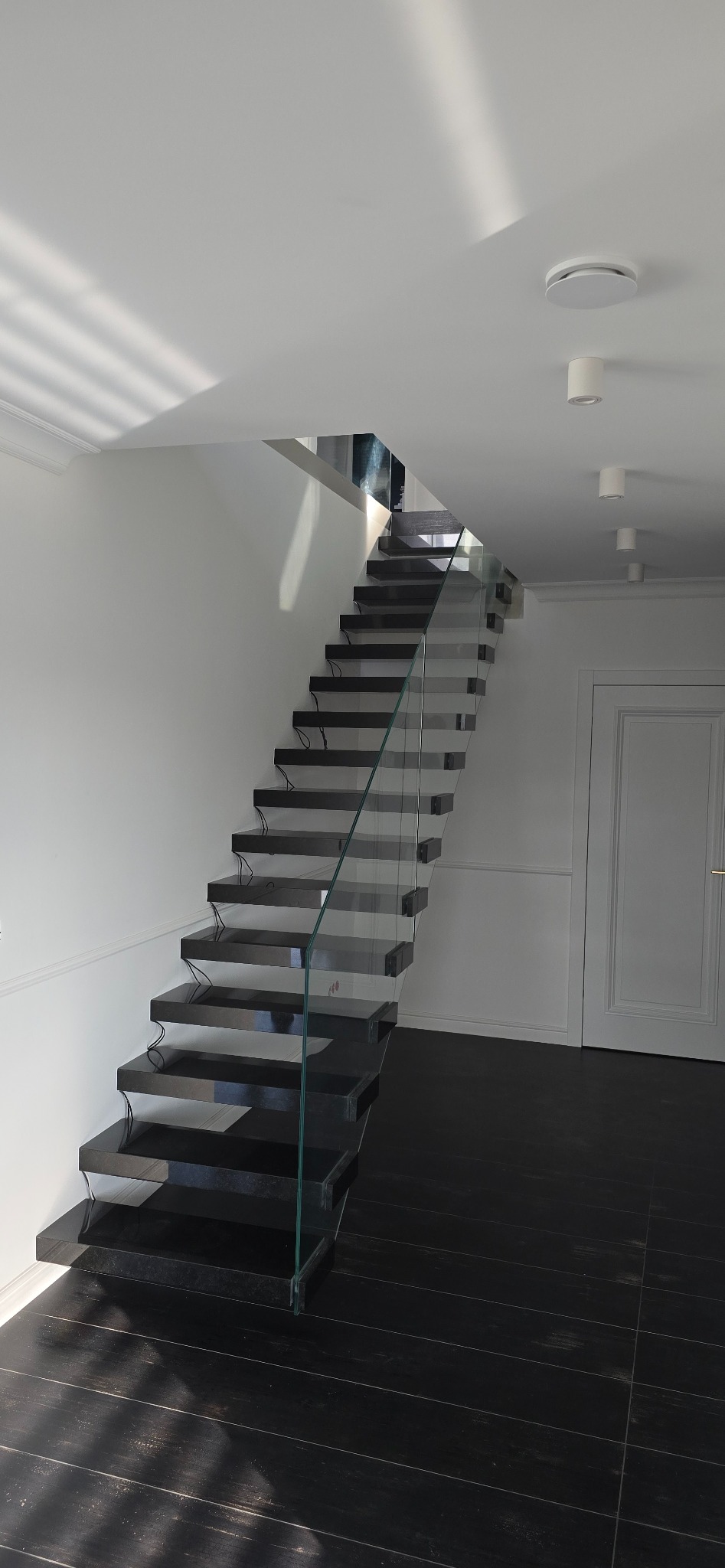 Nowoczesne schody z czarnego kamienia z przezroczystą szklaną balustradą w jasnym wnętrzu z białymi ścianami i ciemną podłogą.