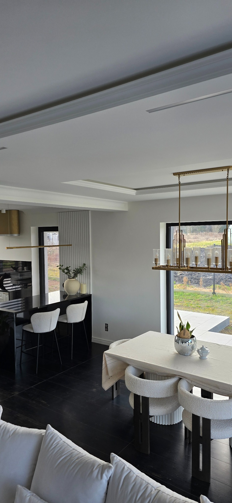 Nowoczesne wnętrze: czarna kuchnia z białymi hokerami, stół z kremowym obrusem i krzesłami typu boucle, złoty żyrandol, widok na ogród przez duże okno.