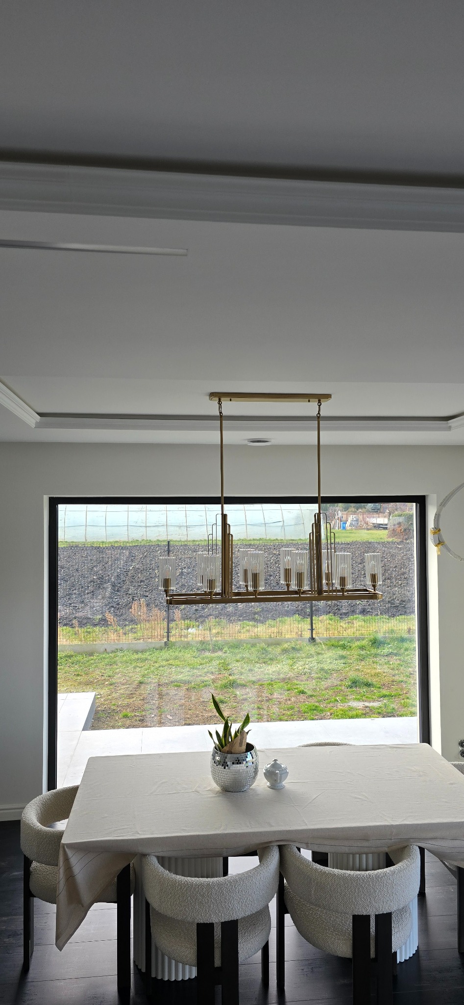 Jadalnia z prostokątnym stołem nakrytym białym obrusem, otoczona krzesłami z tapicerką typu baranek, nad stołem złoty, podłużny żyrandol z szklanymi kloszami, za oknem widok na ogród z siatką...