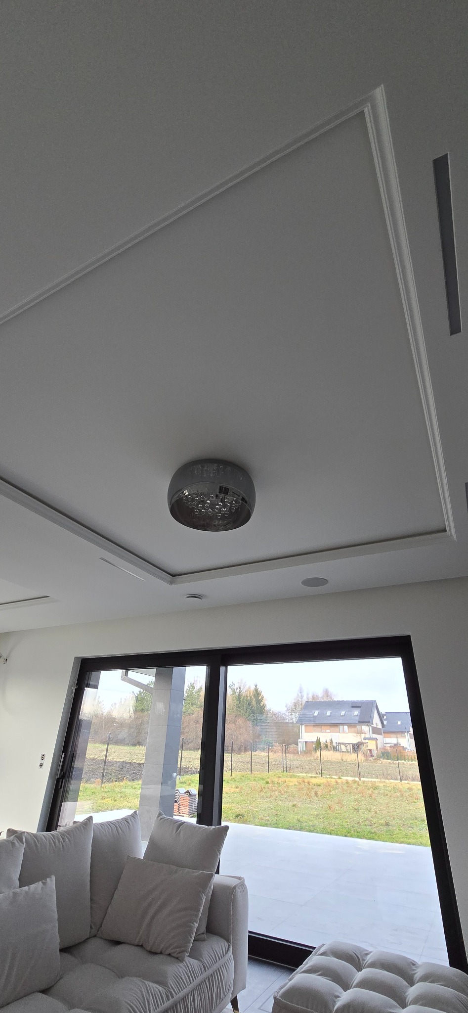 Nowoczesny salon z białymi ścianami, dekoracyjnym sufitem podwieszanym z oświetleniem punktowym i dużym oknem panoramicznym z widokiem na zewnątrz.