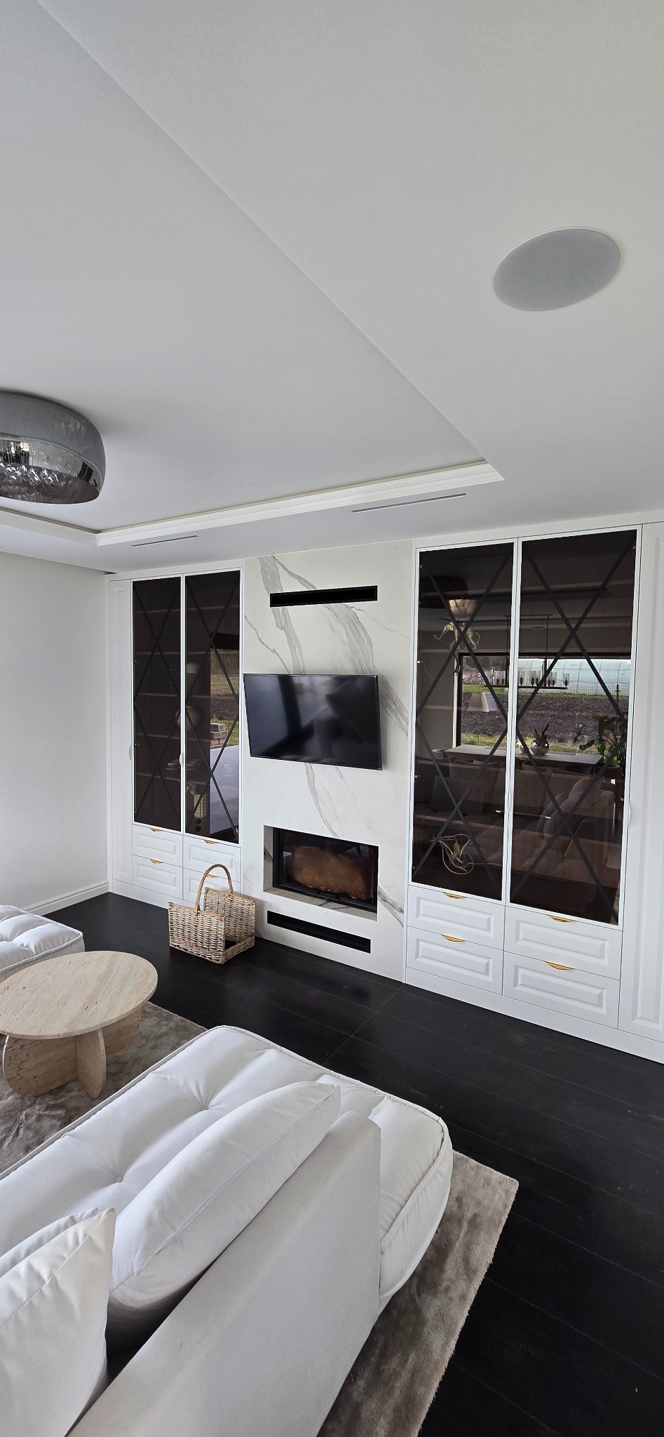 Nowoczesny salon z zabudową meblową w kolorze białym, z marmurowym panelem, w którym umieszczono telewizor i biokominek, oraz lustrzanymi drzwiami szafek z geometrycznym wzorem.
