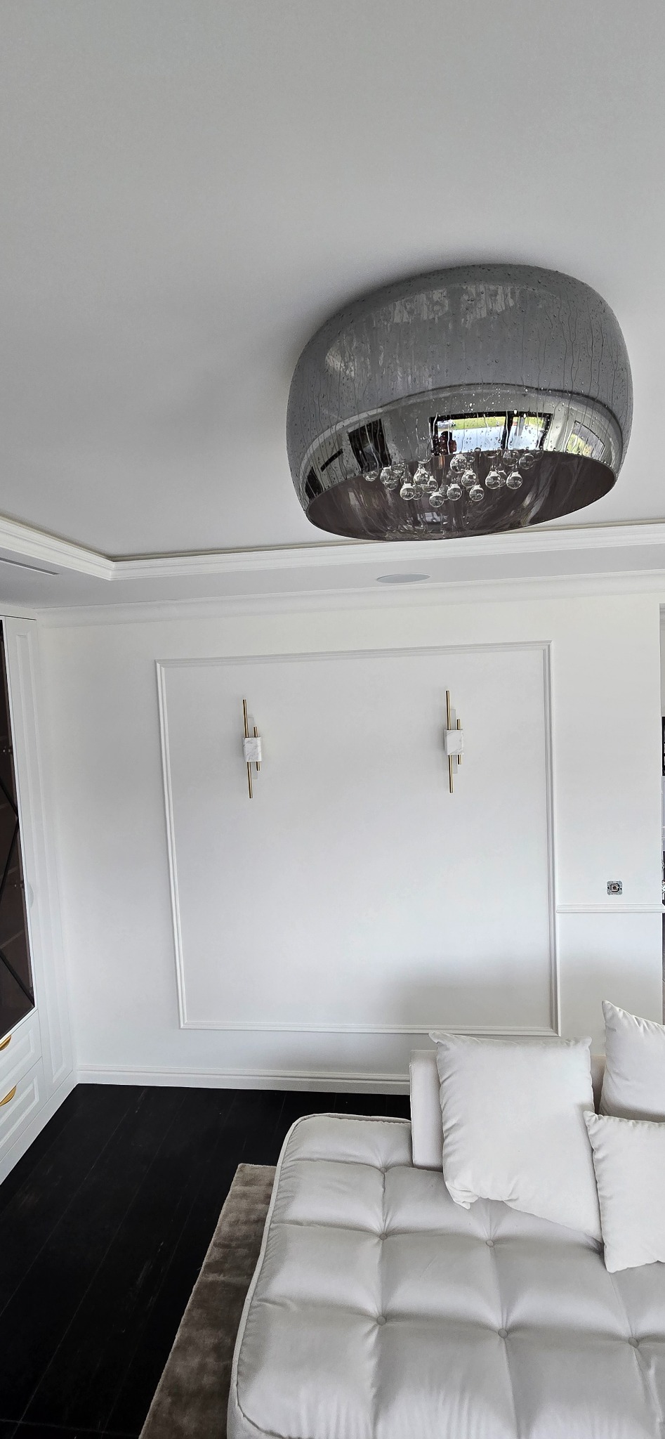 Nowoczesne wnętrze z białymi ścianami z dekoracyjnymi listwami, ciemną drewnianą podłogą, białą sofą i okrągłą lampą sufitową z elementami szklanymi.