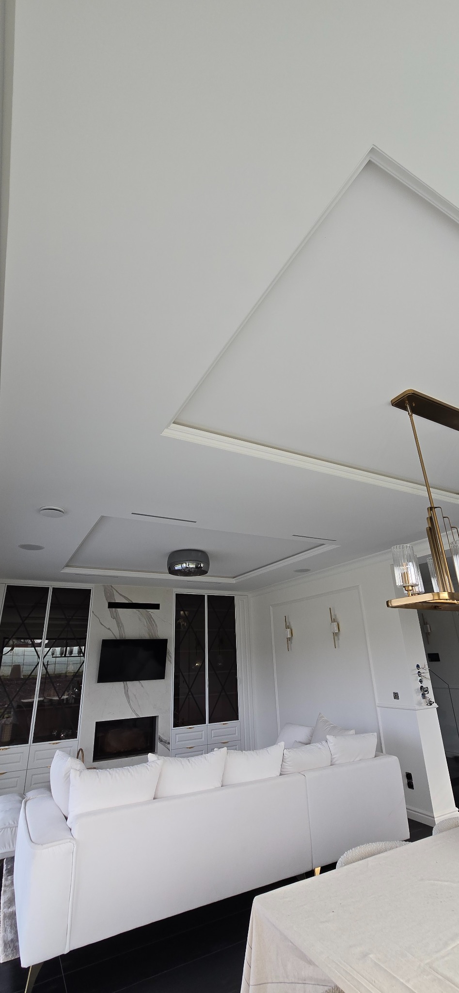 Białe wnętrze salonu z narożną sofą, zabudową meblową z czarnymi wstawkami i marmurowym kominkiem, oświetlone złotą lampą wiszącą i punktowymi lampami ściennymi.