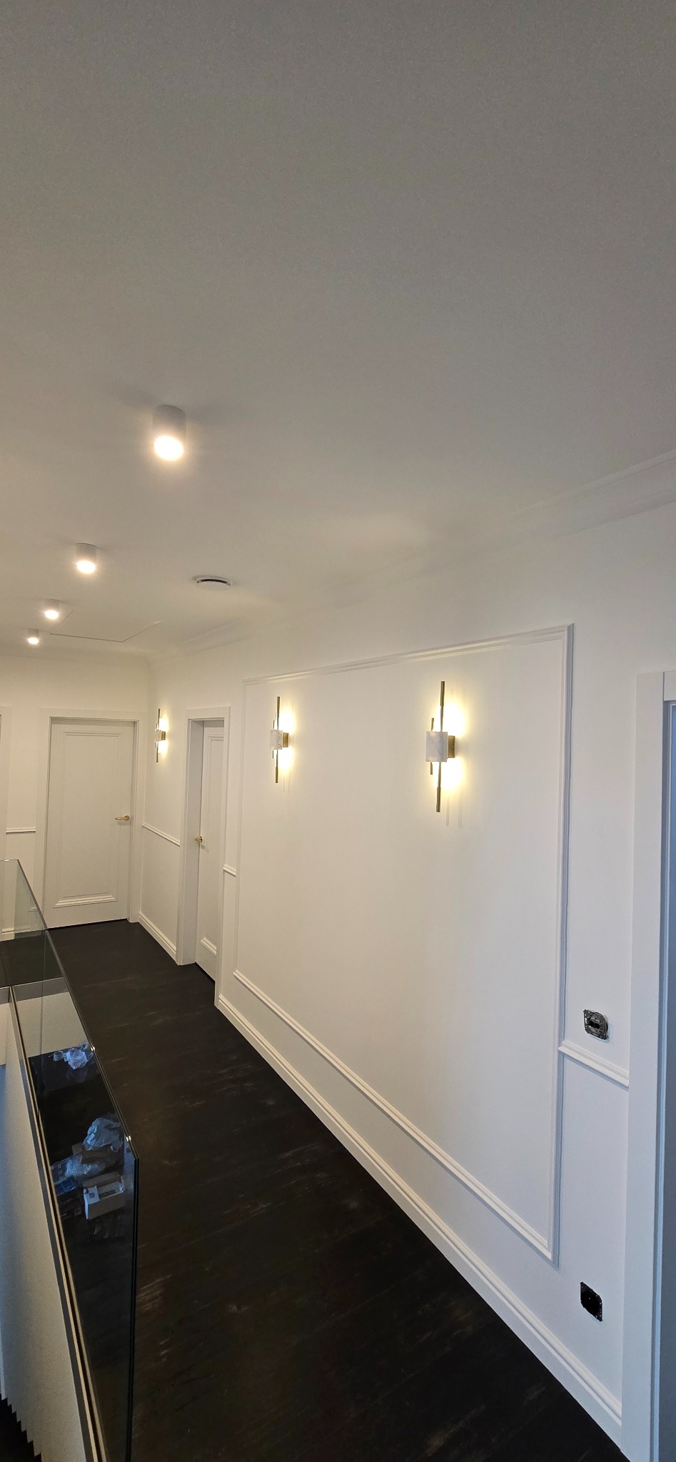 Korytarz po remoncie z ciemną podłogą, białymi ścianami z dekoracyjnymi listwami i nowoczesnymi złotymi kinkietami.
