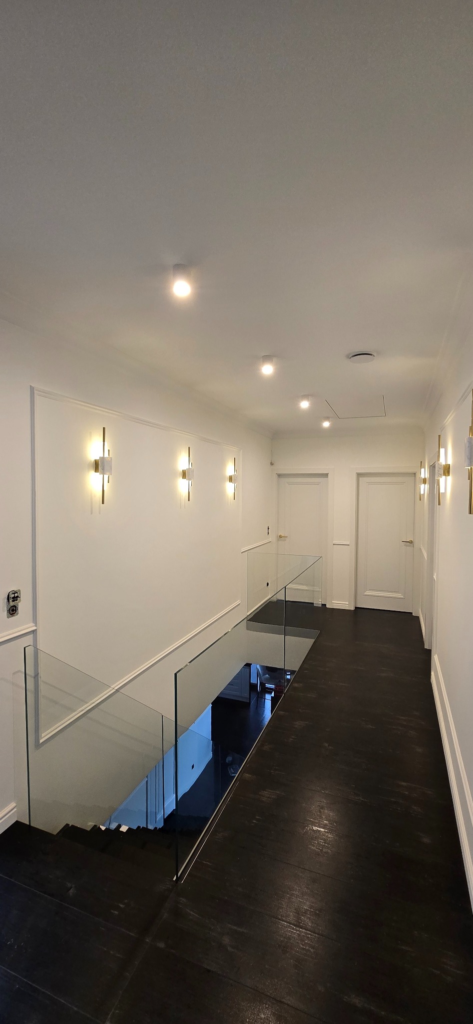 Nowoczesny korytarz z czarną podłogą, szklaną balustradą schodów i białymi ścianami z dekoracyjnymi listwami oraz złotymi kinkietami