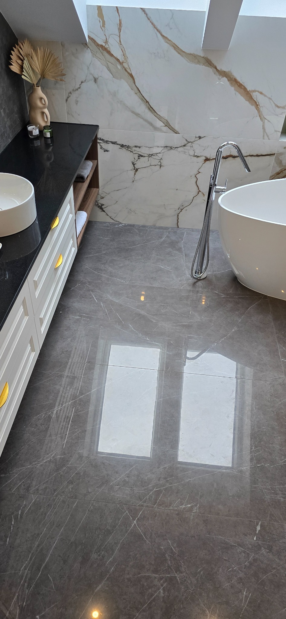 Elegancka łazienka z szarą marmurową podłogą, białą wolnostojącą wanną i umywalką nablatową na czarnym blacie z białymi szafkami, akcentowana złotymi uchwytami i dekoracyjną trawą pampasową w wazonie.