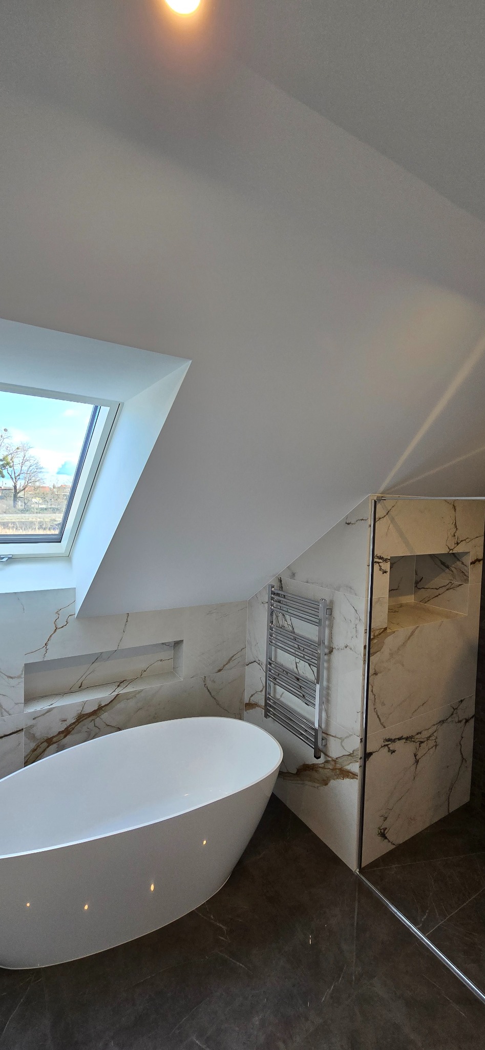 Nowoczesna łazienka na poddaszu z owalną wanną, grzejnikiem drabinkowym i kabiną prysznicową wykończoną marmurowymi płytkami z żyłkowaniem, doświetlona oknem dachowym.