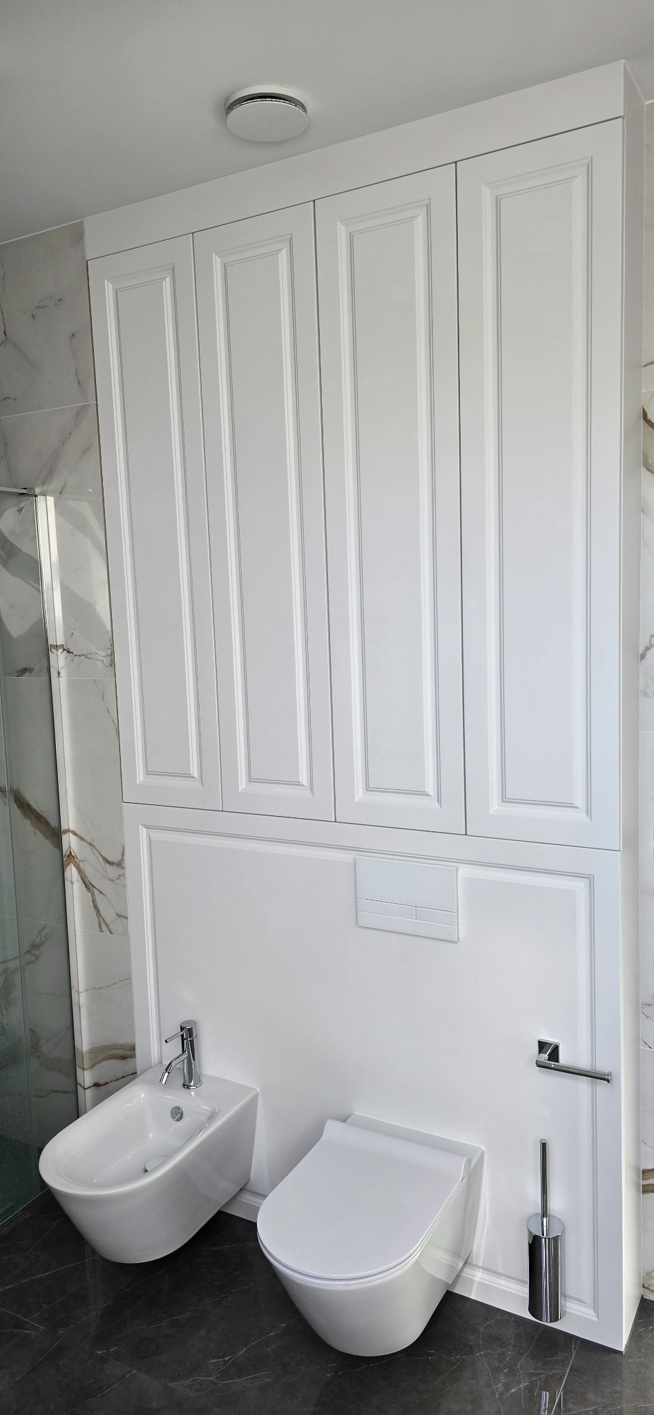 Nowoczesna łazienka z białymi szafkami zabudowanymi nad toaletą i bidetem, marmurowe płytki na ścianach, ciemna posadzka.