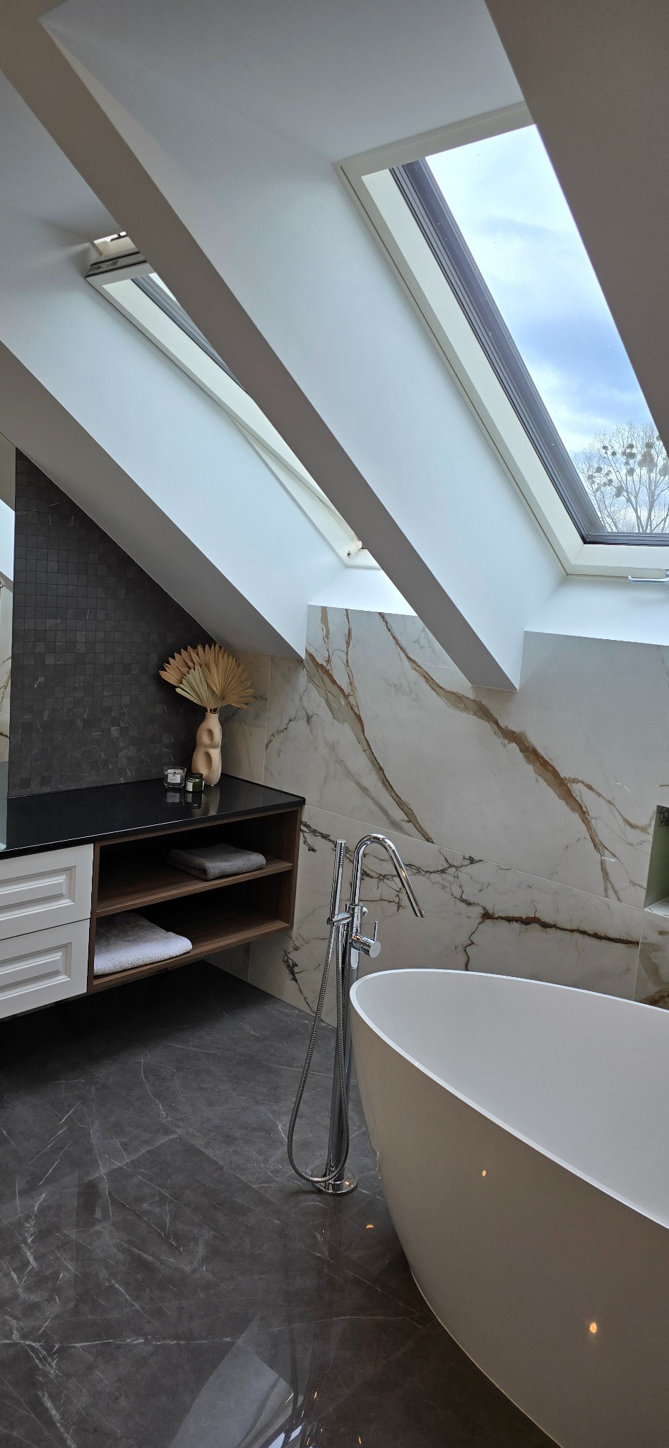 Nowoczesna łazienka na poddaszu z wanną wolnostojącą, marmurowymi płytkami ściennymi z brązowymi żyłami, szarą podłogą z marmuru, szafką z czarnym blatem i drewnianymi półkami, oraz oknami dachowymi.