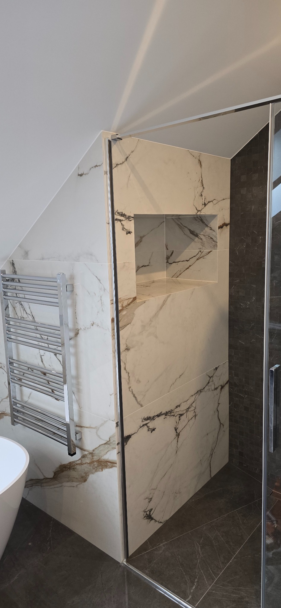 Nowoczesna kabina prysznicowa z marmurowymi płytkami w odcieniach bieli i szarości, wbudowana półka w ścianie, grzejnik drabinkowy, skośny sufit.