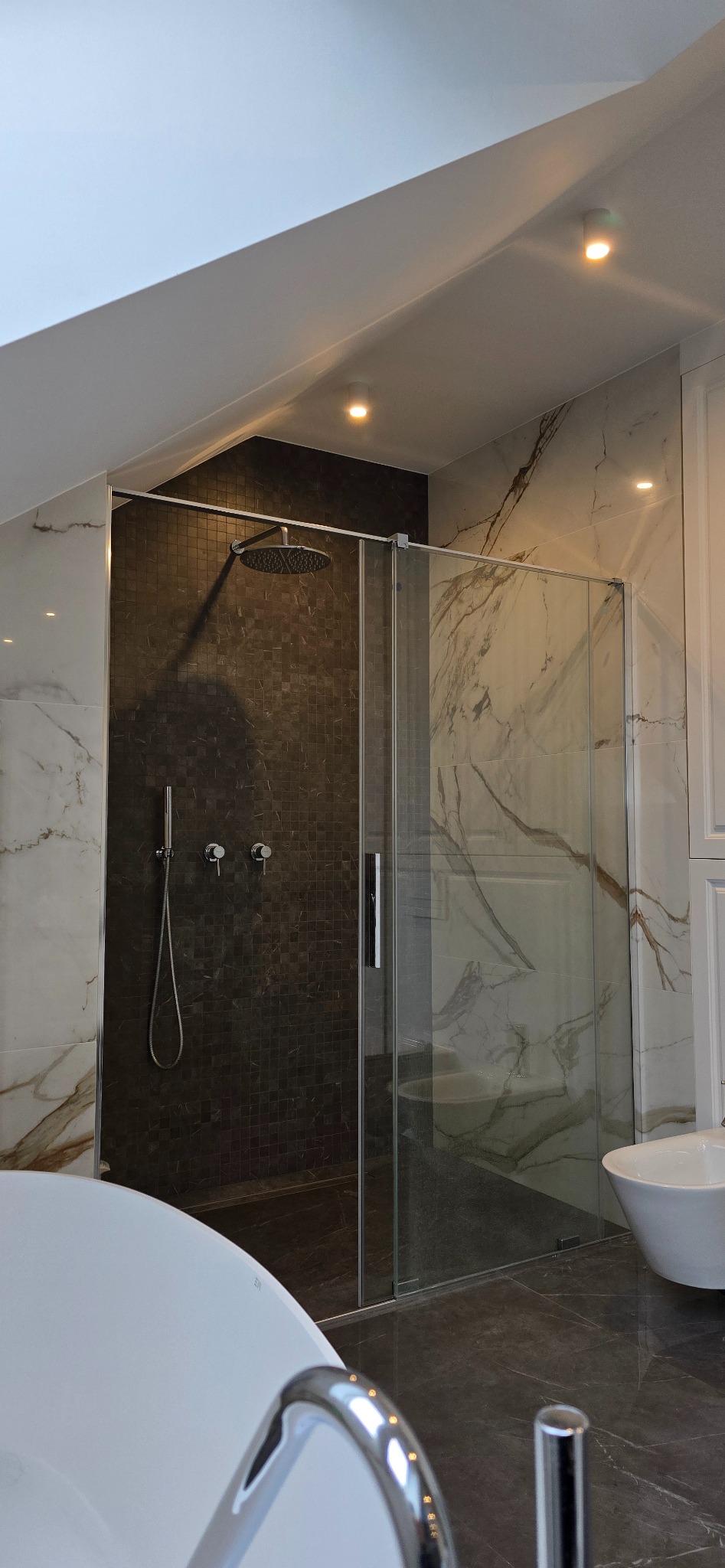 Nowoczesna łazienka z kabiną prysznicową typu walk-in, wykończona płytkami imitującymi marmur i mozaiką, oświetlona punktowymi lampami sufitowymi.