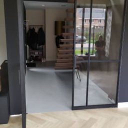 Luxury Raw Design - Nowoczesne wnętrze z szerokim, czarnym, przesuwnym systemem szklanych drzwi, widok na drewniane schody spiralne i szarą podłogę, parkiet ułożony w jodełkę.