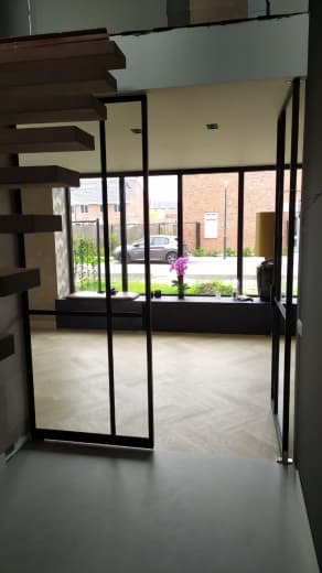 Nowoczesne, czarne drzwi przesuwne z metalową ramą, widok z wnętrza domu na ogród i ulicę, podłoga w jasny wzór jodełki, schody drewniane po lewej stronie.