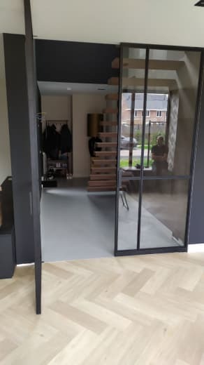 Nowoczesne wnętrze z szerokim, czarnym, przesuwnym systemem szklanych drzwi, widok na drewniane schody spiralne i szarą podłogę, parkiet ułożony w jodełkę.