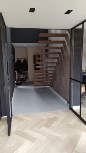 Nowoczesne wnętrze z otwartymi schodami z drewna, szarą podłogą i parkietem w jodełkę, widoczne czarne ramy drzwi.