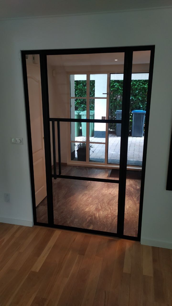 Nowoczesne drzwi przesuwne z czarną ramą i szklanymi panelami, widok na taras z zielenią i pojemnikami na śmieci, drewniana podłoga wewnątrz.