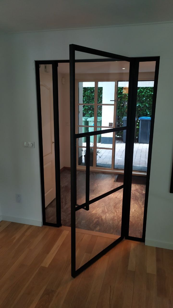 Częściowo otwarte, nowoczesne drzwi z czarną ramą i szklanymi panelami, widok na wnętrze domu i ogród z kostką brukową oraz roślinnością.