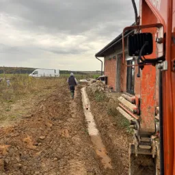 Wykopany rów pod instalację, widoczny fragment pomarańczowej koparki Hitachi, pracownik w oddali i budynek w trakcie budowy w tle, pochmurne niebo.