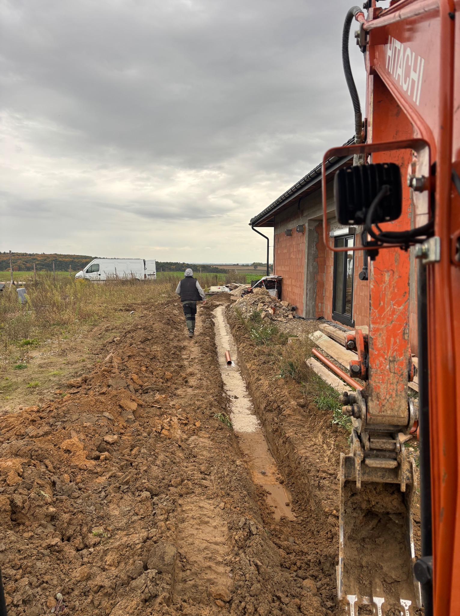 Wykopany rów pod instalację, widoczny fragment pomarańczowej koparki Hitachi, pracownik w oddali i budynek w trakcie budowy w tle, pochmurne niebo.