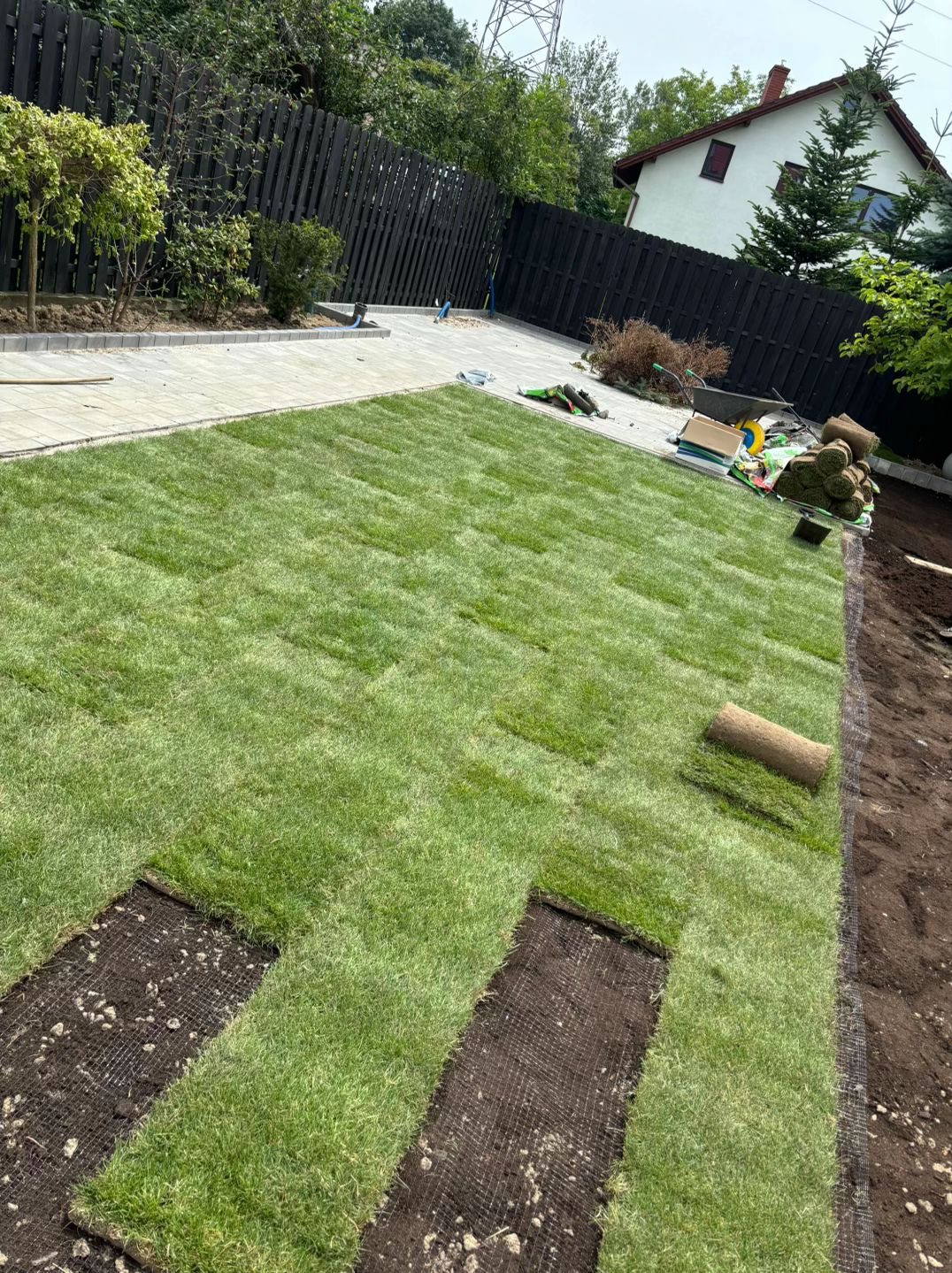 Układanie trawy z rolki w ogrodzie. Widoczne fragmenty darni, rolki trawy, przygotowane podłoże i narzędzia ogrodnicze. W tle dom i ogrodzenie.