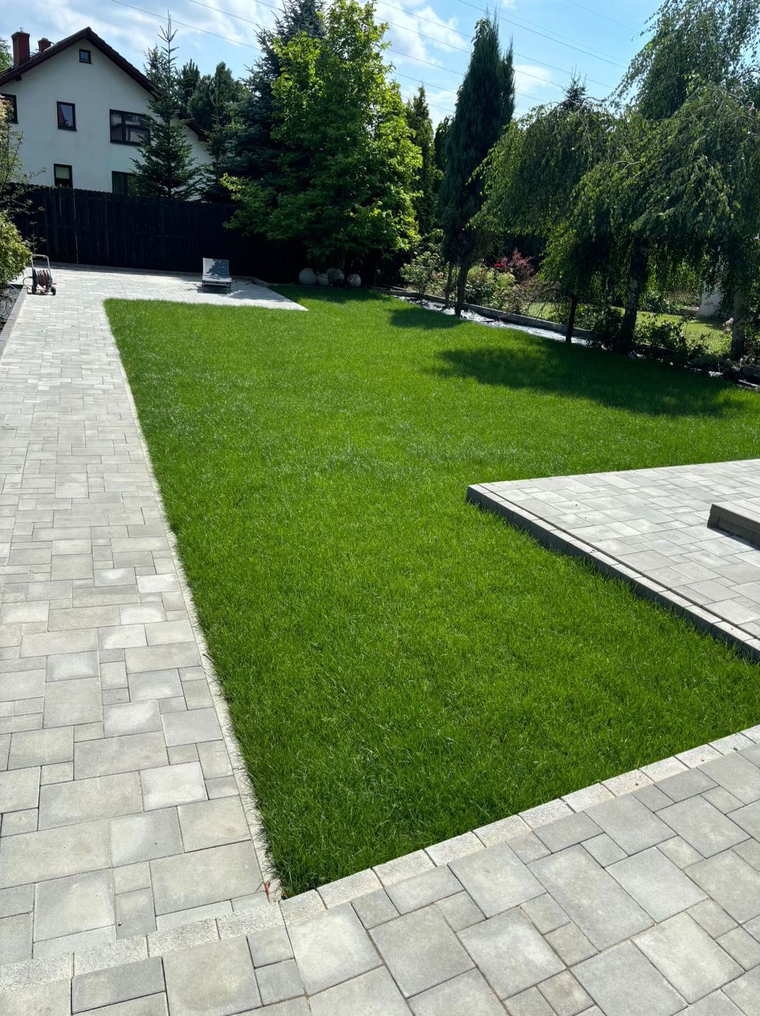 Zadbany trawnik z równo przystrzyżoną trawą, obrzeża z kostki brukowej, drzewa w tle. Widok na wypielęgnowany ogród w słoneczny dzień.
