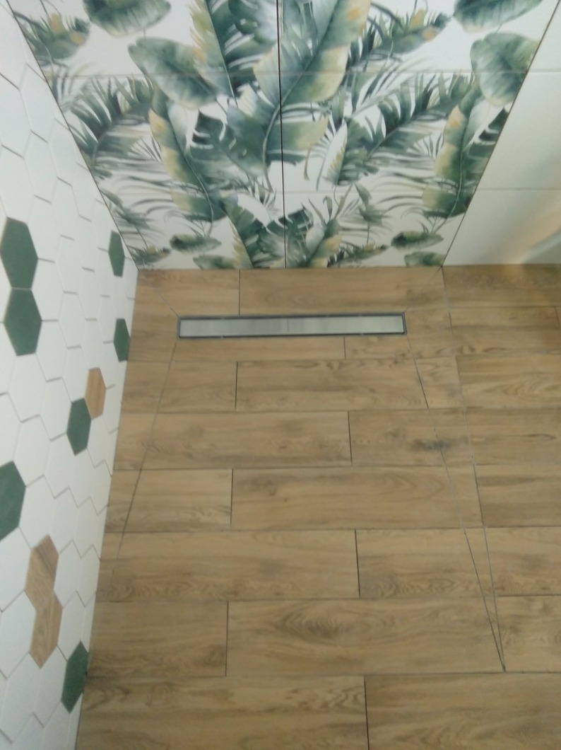 Wykończona łazienka z drewnopodobnymi płytkami podłogowymi, odpływem liniowym, heksagonalnymi płytkami ściennymi w odcieniach bieli, zieleni i brązu oraz płytkami na suficie z motywem tropikalnych...