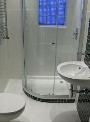 Biała łazienka z kabiną prysznicową z zaokrąglonym narożnikiem, umywalką i toaletą. W ścianie okno z luksferów w kolorze niebieskim.