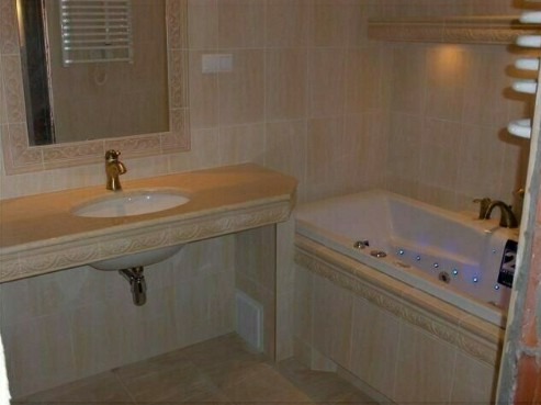 Jasna łazienka z wanną z hydromasażem i umywalką nablatową z baterią w kolorze złotym, wykończona beżowymi płytkami z dekoracyjnym frezem.