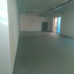 Wnętrze pomieszczenia w stanie surowym z wylaną posadzką jastrychową, widoczne rury pod sufitem i przygotowane ściany do dalszych prac wykończeniowych.