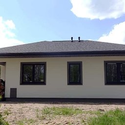 AK.MK Bud - Wykończony dom jednorodzinny z szarą dachówką i beżową elewacją, widok z boku na tle lasu i błękitnego nieba z chmurami.