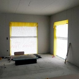 AK.MK Bud - Wnętrze w trakcie prac wykończeniowych: ściany obłożone płytami gipsowo-kartonowymi, okna zabezpieczone folią, widoczne narzędzia i materiały budowlane na podłodze.