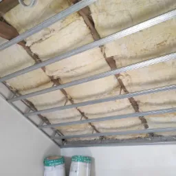 Montaż izolacji z wełny mineralnej w przestrzeni pod stropem, widoczne metalowe profile konstrukcyjne i rolki wełny Platinum 32 w tle.