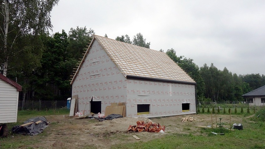 Budynek w trakcie budowy, zabezpieczony folią, z widoczną konstrukcją dachu, przygotowany do dalszych prac elewacyjnych.