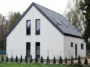 Nowoczesny dom jednorodzinny z białą elewacją, grafitowym dachem i oknami, otoczony niskimi tujami i siatką ogrodzeniową.