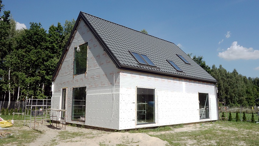 Dom w trakcie budowy, zabezpieczony folią wiatroizolacyjną, widoczne okna i dach z dachówką, rusztowanie obok budynku.