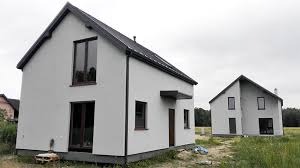 Dwa nowoczesne, pomalowane na szaro domy jednorodzinne z ciemnymi oknami i dachem, stojące na trawiastym terenie w pochmurny dzień. Jeden dom zadaszony nad wejściem.