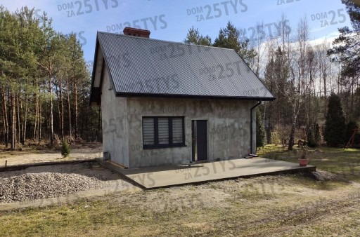 Surowy, nieotynkowany dom jednorodzinny z szarą blachą falistą na dachu, ciemnymi oknami i drzwiami, otoczony lasem i placem z kamieniami.