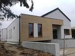 Nowoczesny dom jednorodzinny z elewacją w kolorze białym i elementami z jasnego drewna, widoczne okna i fragment dachu, teren wokół domu w trakcie prac wykończeniowych.