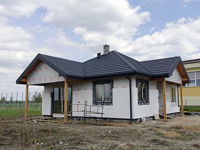Dom jednorodzinny w trakcie ocieplania, widoczne płyty styropianowe na ścianach, czarna dachówka i drewniane słupy werandy.