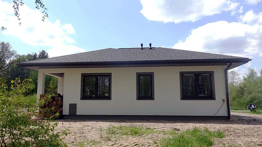 Wykończony dom jednorodzinny z szarą dachówką i beżową elewacją, widok z boku na tle lasu i błękitnego nieba z chmurami.