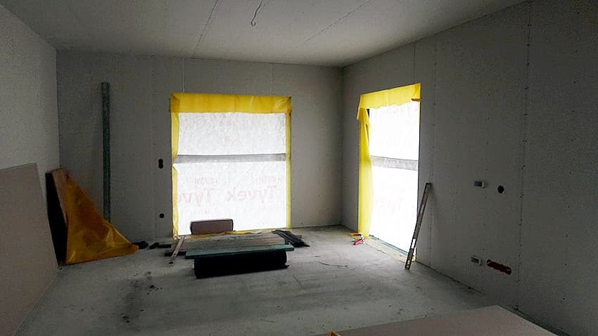 Wnętrze w trakcie prac wykończeniowych: ściany obłożone płytami gipsowo-kartonowymi, okna zabezpieczone folią, widoczne narzędzia i materiały budowlane na podłodze.