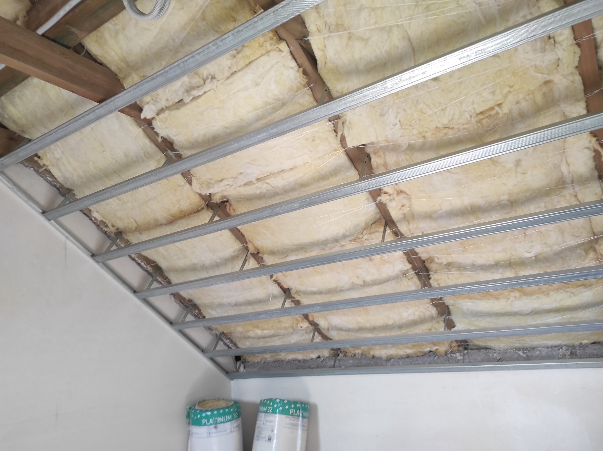 Montaż izolacji z wełny mineralnej w przestrzeni pod stropem, widoczne metalowe profile konstrukcyjne i rolki wełny Platinum 32 w tle.
