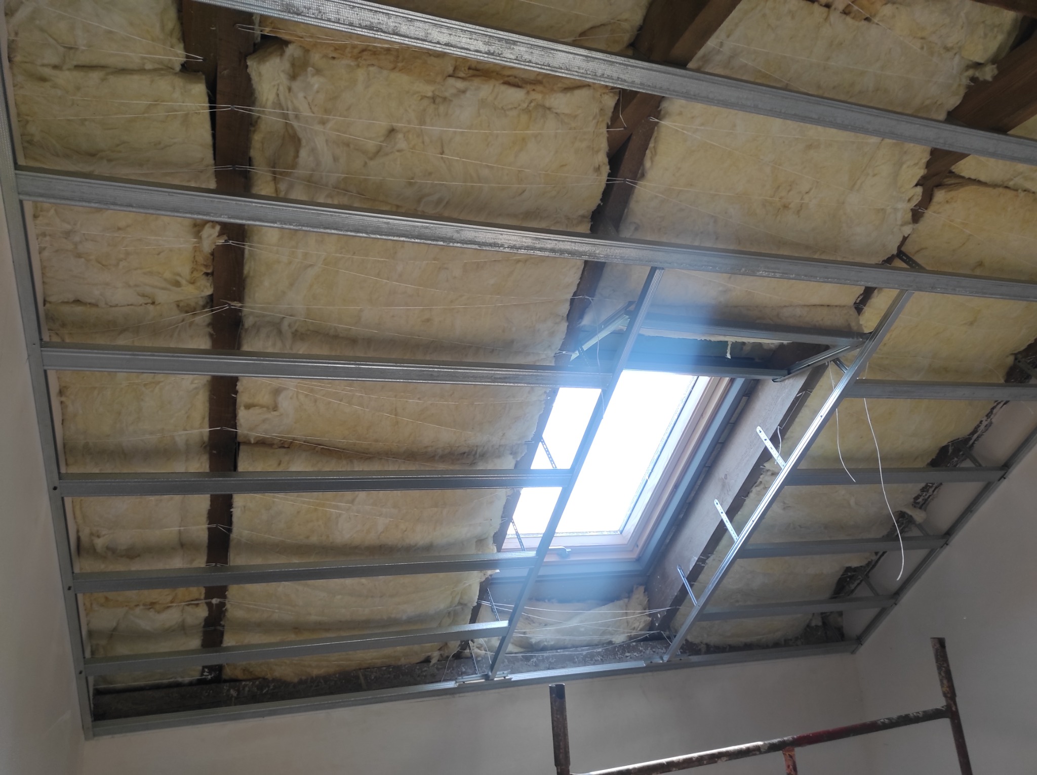 Metalowa konstrukcja sufitu podwieszanego z izolacją z wełny mineralnej i oknem dachowym, widok od dołu.