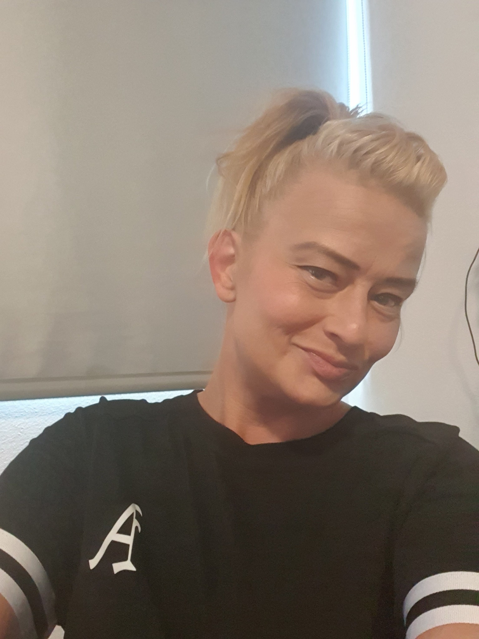 Portret uśmiechniętej blondynki z włosami upiętymi w wysoki kucyk, ubrana w czarną koszulkę z białym logo litery A i białymi paskami na rękawach, na tle jasnej ściany i rolet.