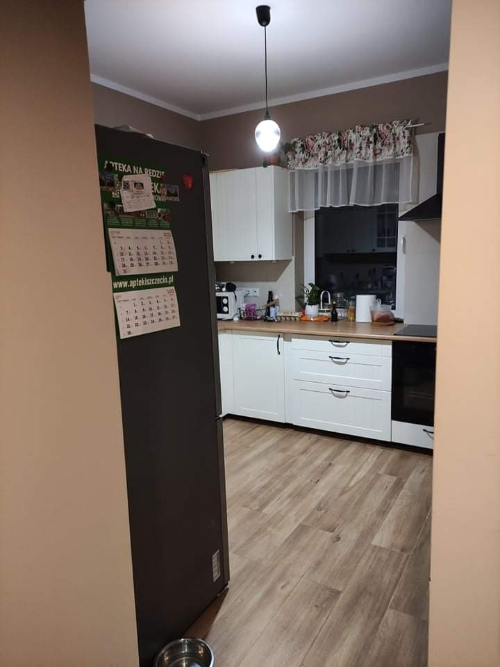 Widok na uporządkowaną kuchnię z drewnopodobną podłogą, białymi szafkami i lodówką z kalendarzem, widziany zza framugi drzwi.