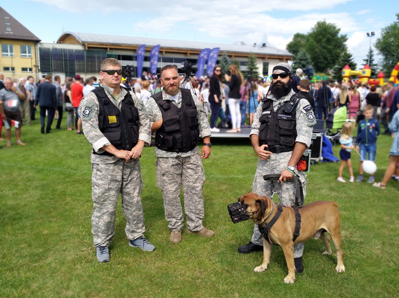 Trzech mężczyzn w mundurach ochronnych i kamizelkach taktycznych stoi na trawie z psem w kagańcu na tle tłumu ludzi i budynku.