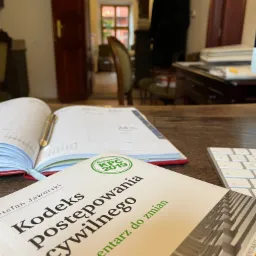 Książka 'Kodeks postępowania cywilnego' leżąca na biurku obok otwartego kalendarza z długopisem i klawiatury, w tle widoczne otwarte drzwi do innego pomieszczenia.