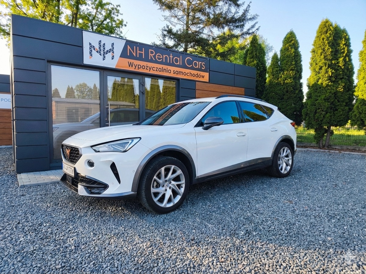 Biały crossover Cupra Formentor przed budynkiem wypożyczalni NH Rental Cars, widok z dołu, szary żwir na podłożu, zieleń drzew w tle.