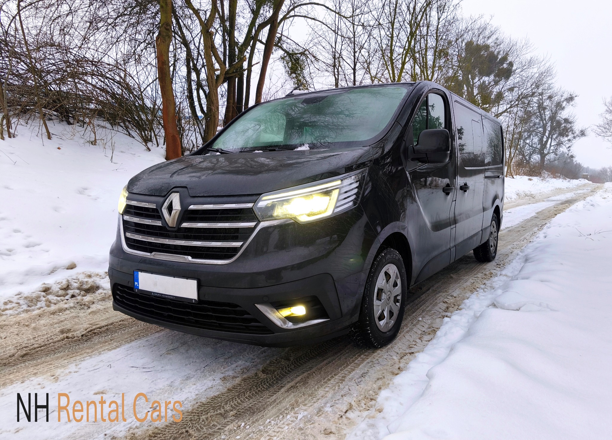 Czarny van Renault Trafic z włączonymi światłami mijania, zaparkowany na ośnieżonej drodze. Widoczne ślady opon w śniegu. W tle drzewa bez liści i logo wypożyczalni.