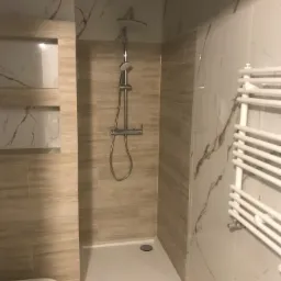 Nowoczesna kabina prysznicowa z deszczownicą i panelem natryskowym, obok grzejnik drabinkowy; ściany wyłożone płytkami imitującymi marmur w odcieniach beżu i bieli, z wnęką.